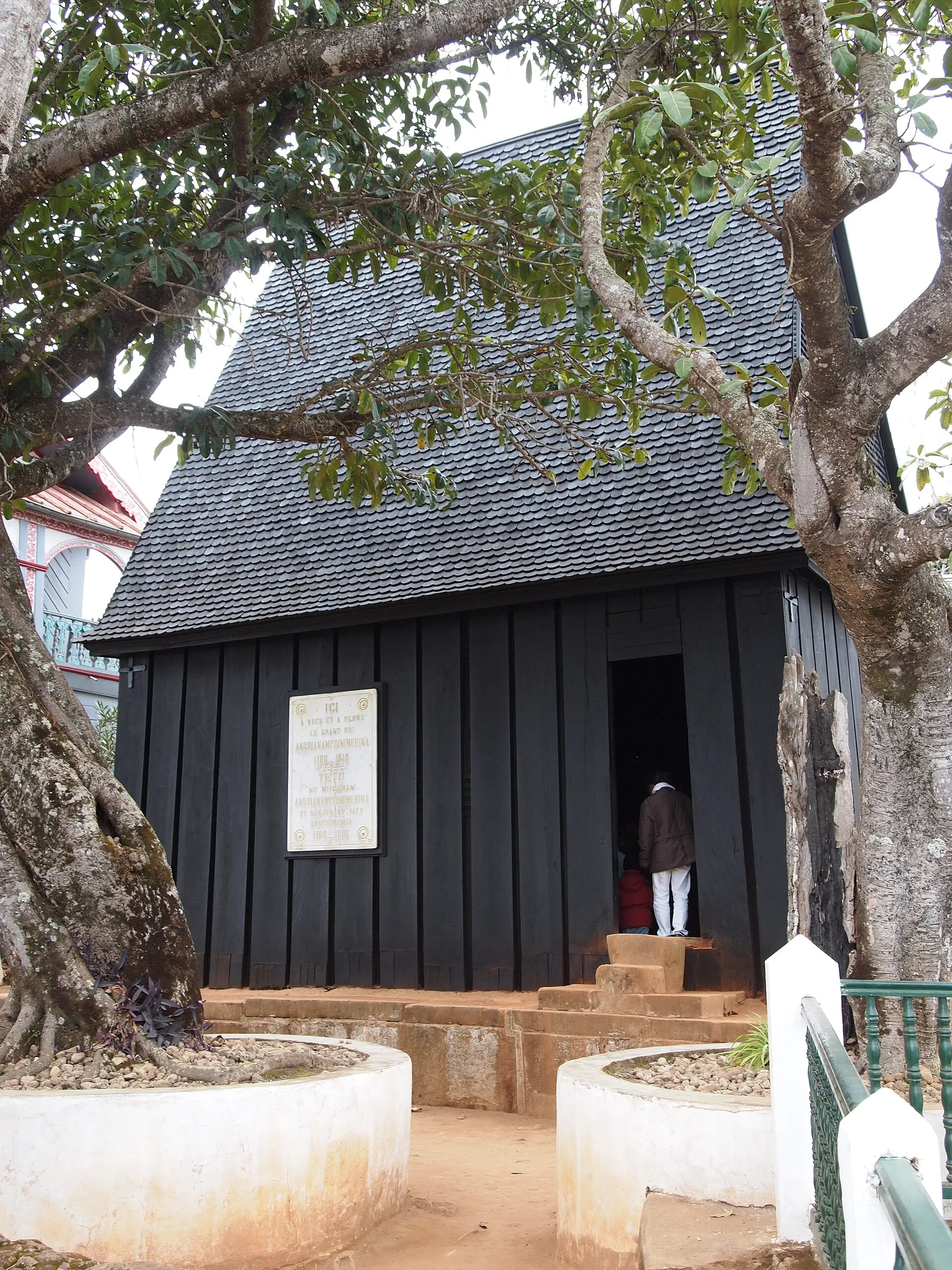 Photographie d'une maison de bois sombre à haut toit de bardeaux, très pentu, dressée sur un socle de pierre au milieu d'arbres. Une porte étroite est ouverte, une personne s'y tient. Un panneau explicatif est fixé sur la façade.