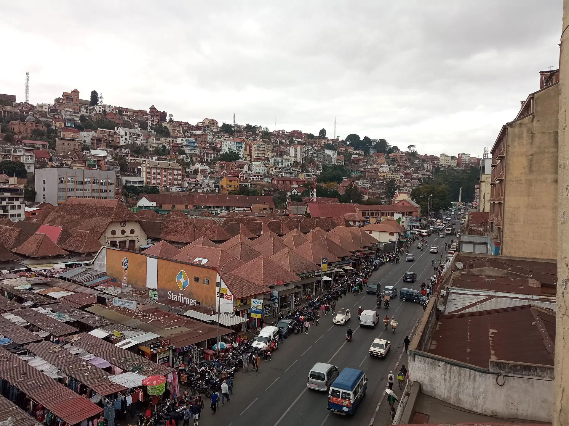 Vue plongeante sur le marché d'Analakely à Antananarivo : les pavillons à toits de tuiles, l'avenue, et la ville qui monte à flanc de colline derrière.