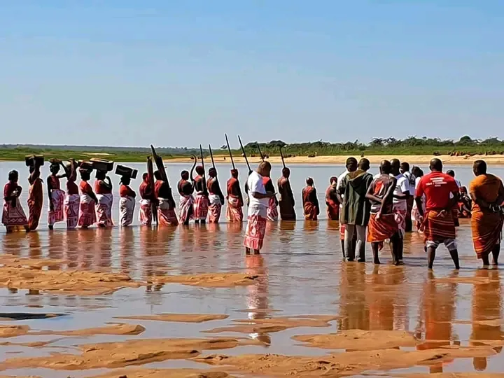 Photographie en couleurs : une foule debout dans le lit peu profond d'un fleuve aux bancs de sable, sous un ciel dégagé. Les participants portent des lambas rouges et à motifs ; plusieurs hampes sont dressées au-dessus des têtes. Une rive boisée ferme l'horizon.