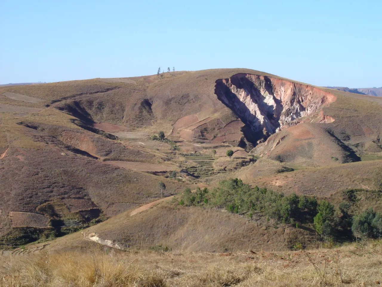 Un lavaka ouvert dans le flanc d'une colline des hautes terres : une entaille claire aux parois abruptes dans un relief de collines pelées.