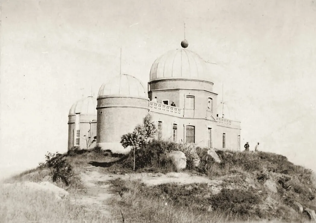Photographie ancienne : l'observatoire d'Ambohidempona sur une colline pelée, trois coupoles de tailles différentes au-dessus d'un bâtiment à balustrade, deux silhouettes minuscules au pied de l'édifice.