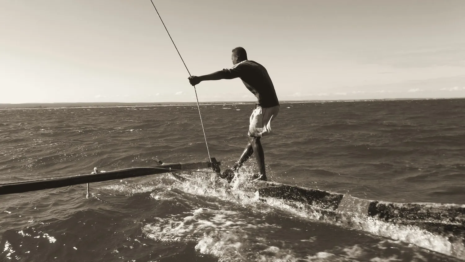 Photographie virée à l'encre : un homme debout sur la coque d'une pirogue lancée, tirant de tout son poids sur l'écoute de la voile, le flotteur du balancier plongeant dans la mer à sa gauche.