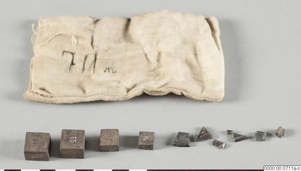Sur fond gris, un petit sac de toile écrue marqué à l'encre « 711 ». Devant lui, alignés, quatre poids de fer cubiques de tailles décroissantes, frappés d'un motif floral, puis neuf petits fragments d'argent aux bords coupés net, dont l'un porte encore des lettres.