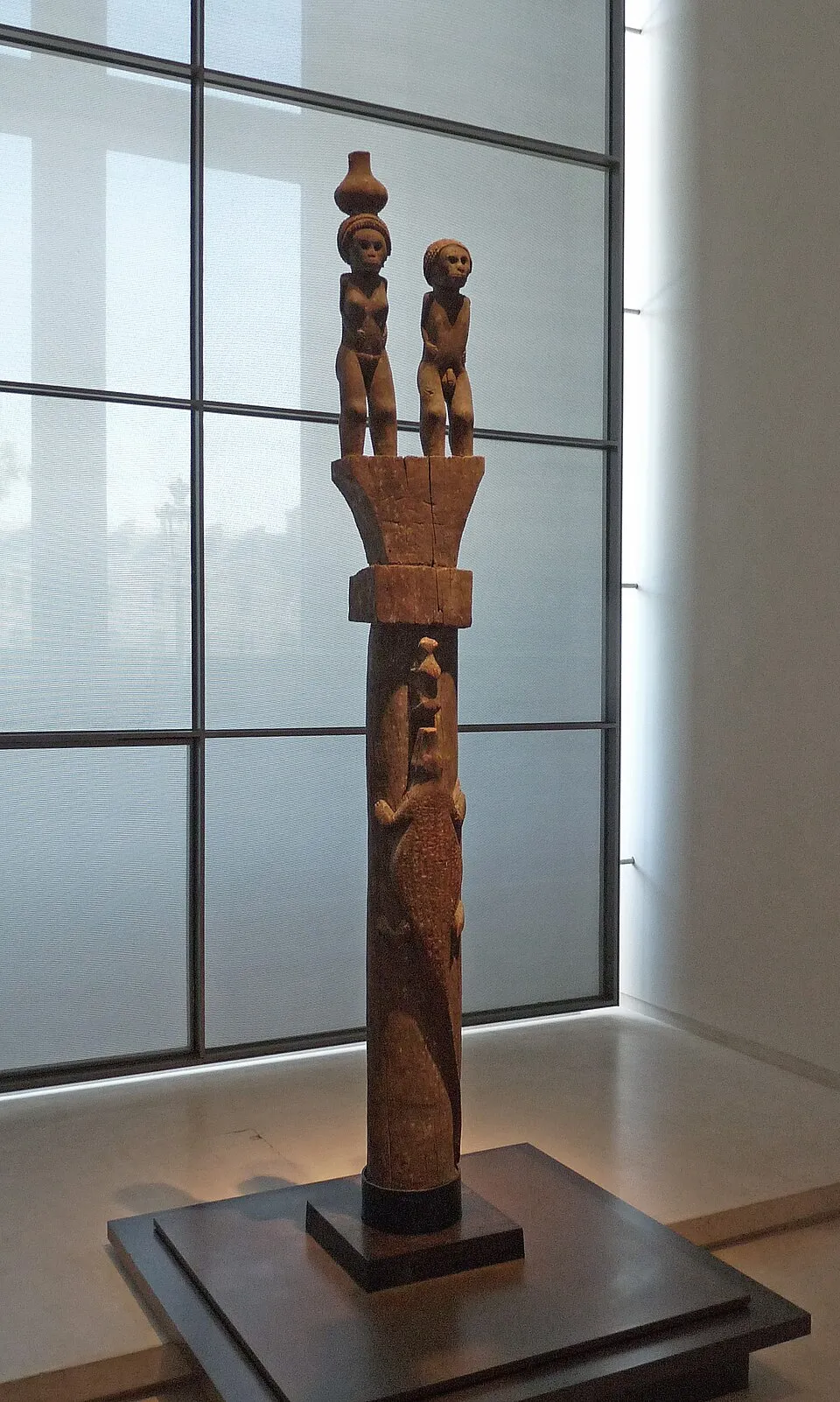 Photographie d'un haut poteau de bois sculpté dressé sur un socle noir dans une salle de musée, devant une paroi vitrée. Le sommet porte deux figures humaines debout et une forme de vase ; le fût est orné d'une figure en relief.