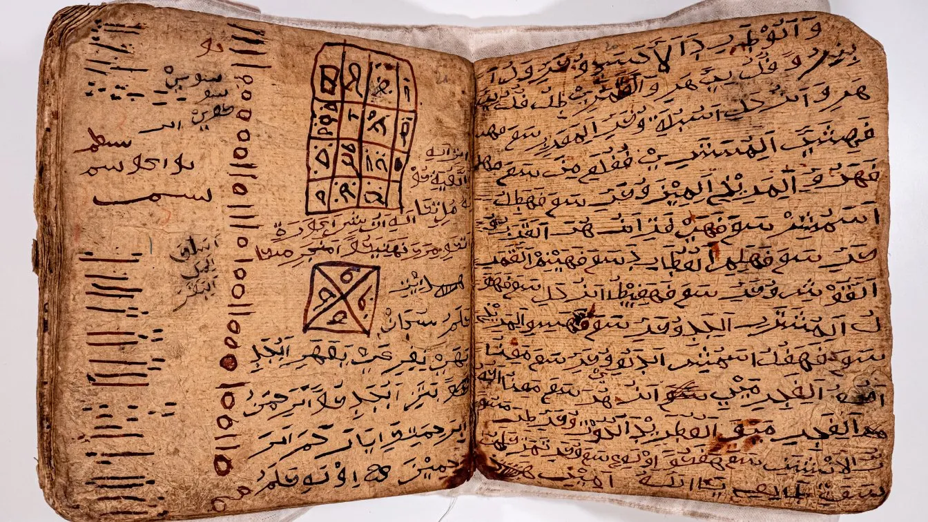 Photographie d'un manuscrit ouvert sur deux pages, posé à plat. Le papier est épais et fibreux, de teinte ocre. Le texte est tracé à l'encre noire et rouge en caractères arabes. La page de gauche porte des colonnes de traits et de points ; la page de droite, en haut, un tableau quadrillé rempli de signes, et plus bas un carré barré d'une croix contenant des figures.