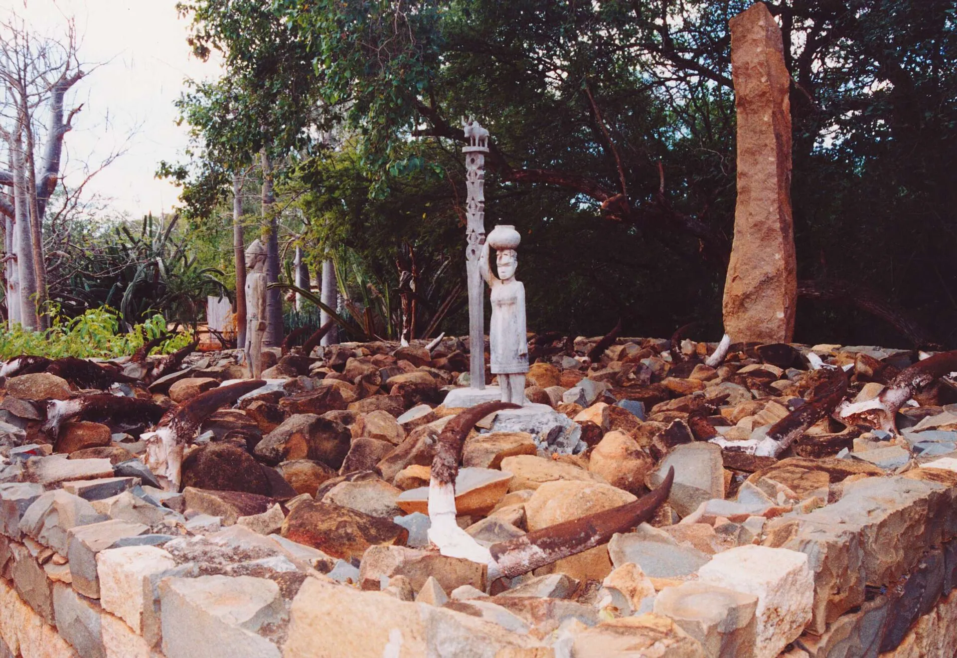 Photographie d'une tombe du Sud malgache : une plateforme rectangulaire de pierres brutes bordée d'un muret bas, hérissée de nombreuses cornes de zébu plantées entre les pierres. Au centre, un poteau sculpté surmonté d'un zébu et une figure de femme portant un vase sur la tête ; à droite, une dalle de pierre dressée. Autour, une végétation sèche d'épineux et de baobabs.