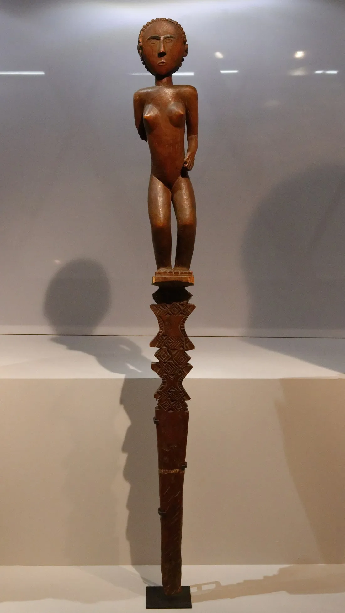 Poteau funéraire en bois dressé, sculpté d'une superposition de figures géométriques ajourées et surmonté d'une silhouette, photographié sur fond neutre en collection de musée.
