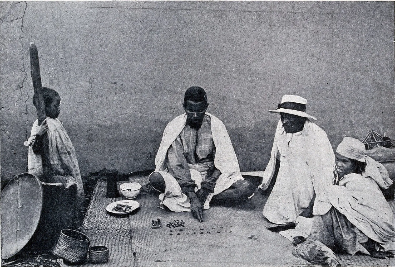 Photographie ancienne : un homme assis en tailleur sur une natte dispose de petits tas de graines en colonnes devant lui ; trois personnes assises autour le regardent faire, parmi un van, une corbeille et un bol.