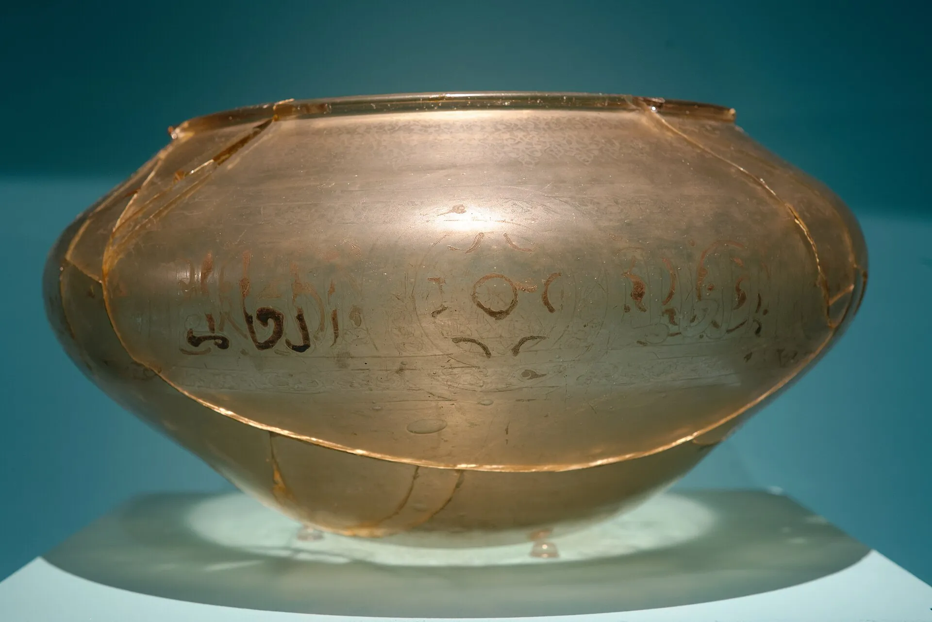 Vase large et bas, translucide et ambré, à panse renflée et lèvre plate, photographié en vitrine sur fond bleu. Un bandeau d'inscription en caractères arabes court sur la panse, encadré de frises gravées plus fines. Des lignes de brisure traversent tout l'objet : il a été cassé puis remonté.